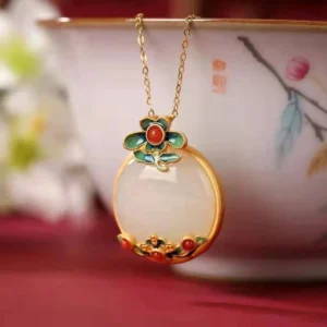White jade flower pendant necklace hung on decorative porcelain teacup, front angle view displaying round milky white jade stone, vibrant red agate accents, refined gold-plated chain, and traditional enamel floral design