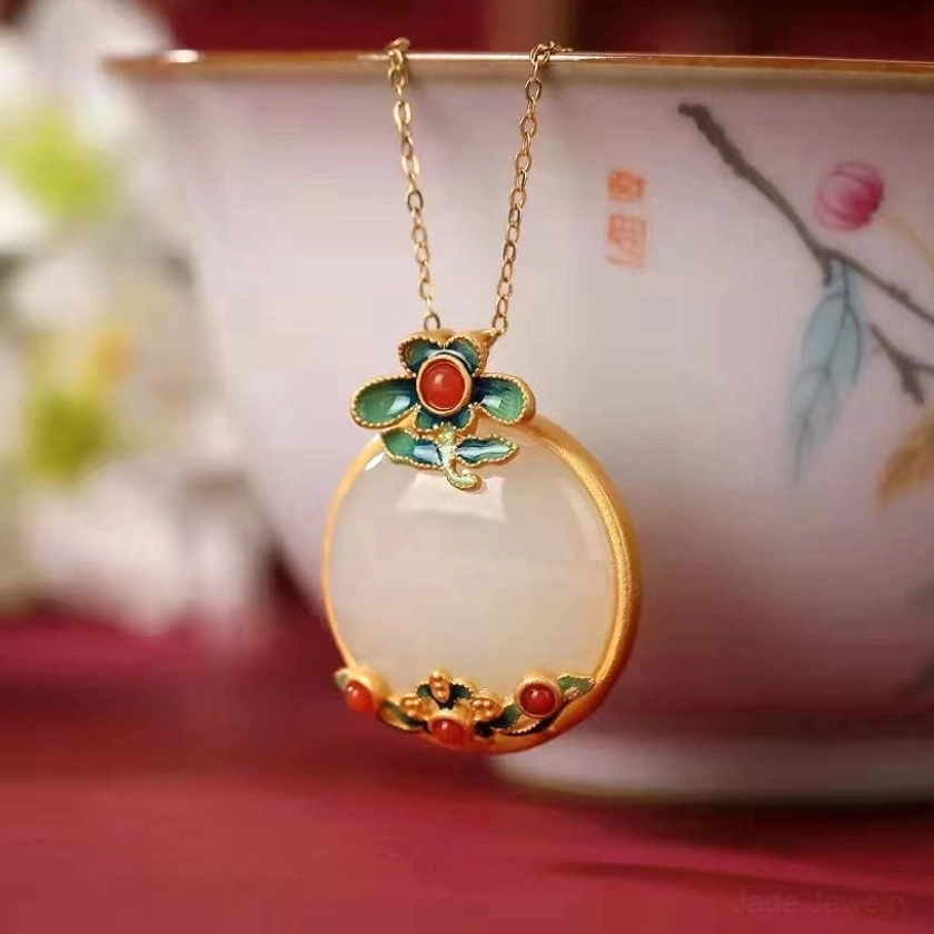 White jade flower pendant necklace hung on decorative porcelain teacup, front angle view displaying round milky white jade stone, vibrant red agate accents, refined gold-plated chain, and traditional enamel floral design